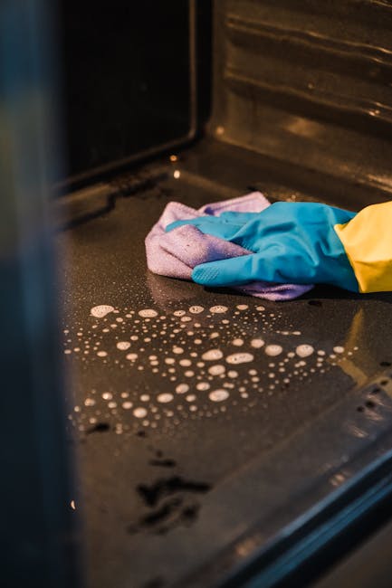 A woman wearing a white t-shirt, yellow and blue rubber gloves, and holding a spray bottle in her right hand, is deep cleaning the interior of a black oven with a purple cloth. The kitchen has white c