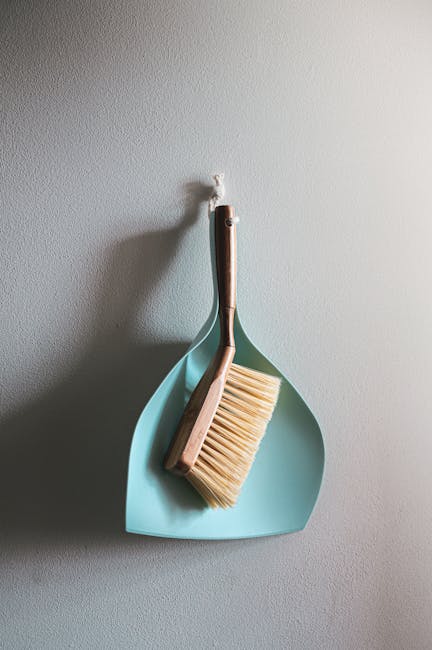 A light blue dustpan hangs on a white textured wall, with a wooden-handled broom resting inside it. The broom features beige bristles and appears clean and well-maintained, positioned in a vertical or