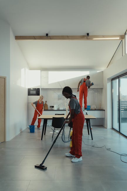 A light blue dustpan hangs on a white textured wall, with a wooden-handled broom resting inside it. The broom features beige bristles and appears clean and well-maintained, positioned in a vertical or