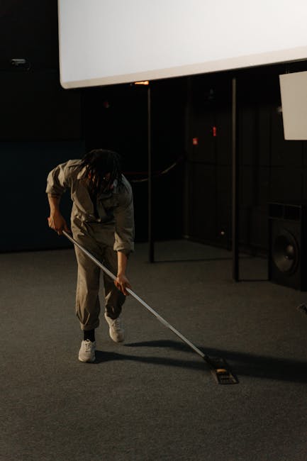 A young boy and an older man are engaged in domestic cleaning in a bright living room. The boy is using a vacuum cleaner on a light-colored fabric sofa, which appears clean and well-maintained. The ma